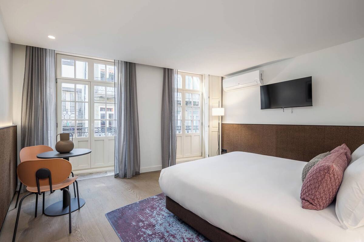Habitación de hotel con cama, escritorio, silla, televisor y ventanas grandes con cortinas.