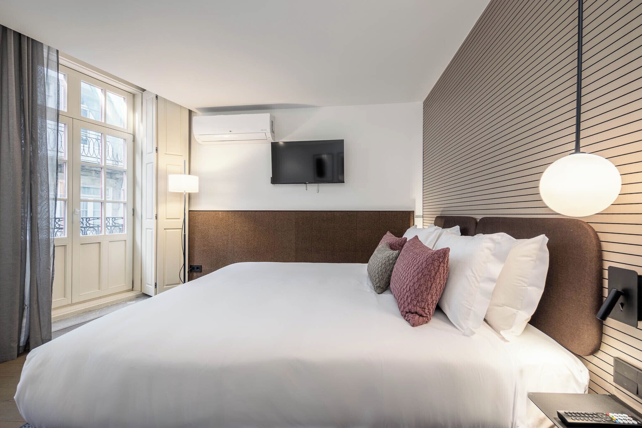 A hotel room with a large bed, a flat-screen TV mounted on the wall, and a window with curtains.