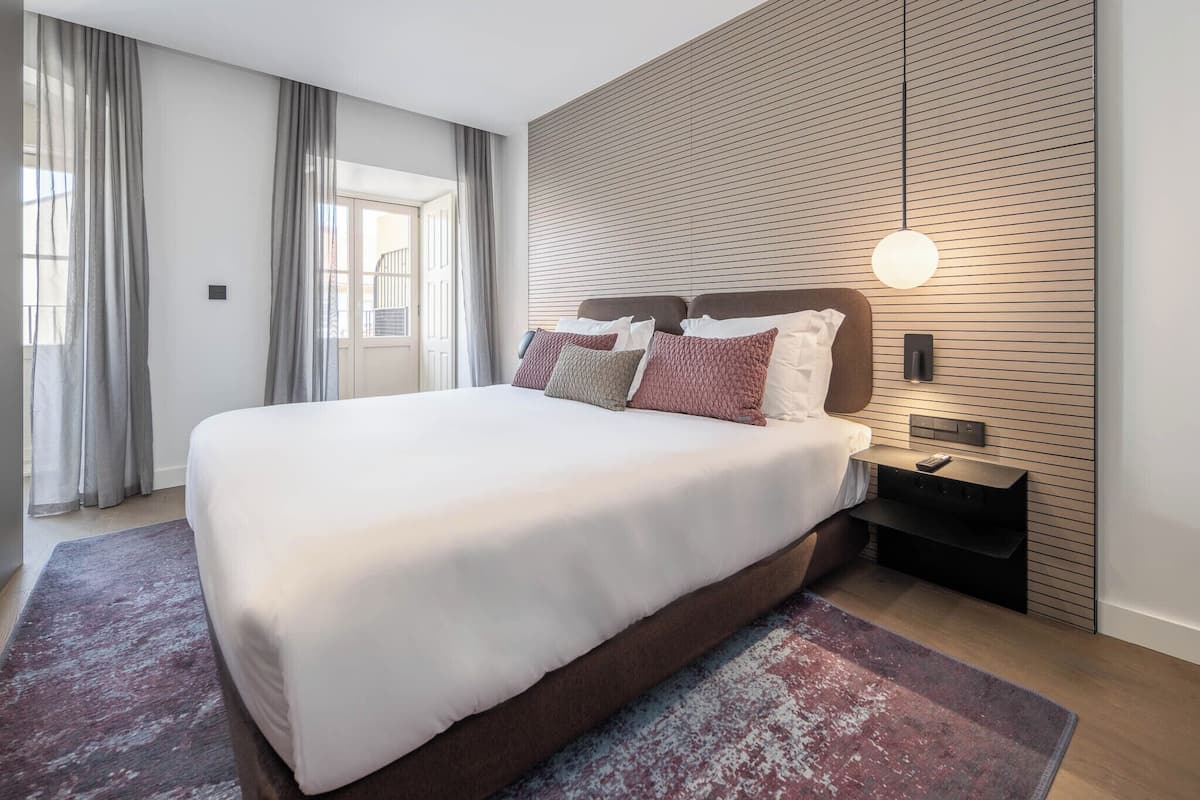 Una habitación de hotel moderna con una cama grande, un panel de pared de madera, una mesita de noche con lámpara y vistas al exterior por la ventana.