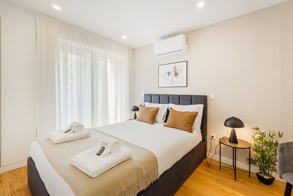 Habitación de hotel con cama, mesita de noche con lámpara, planta en maceta y aire acondicionado.