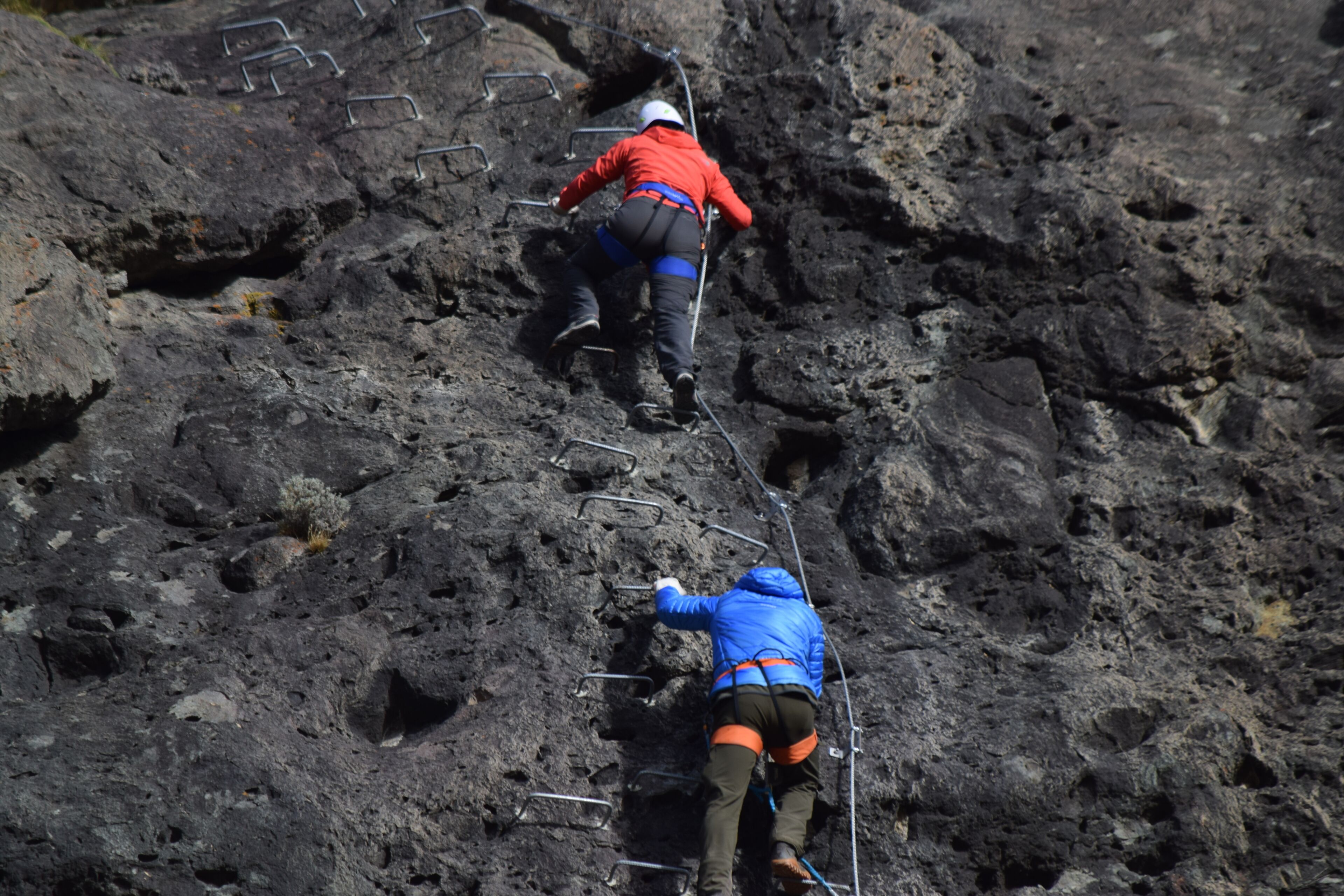Deux personnes escaladent un terrain rocheux à l’aide d’échelons métalliques.