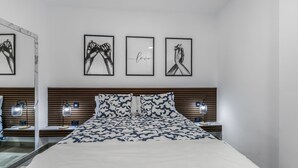 Une chambre moderne avec un grand lit, deux tables de chevet avec des lampes, une tête de lit en bois et trois œuvres d’art encadrées accrochées au mur.