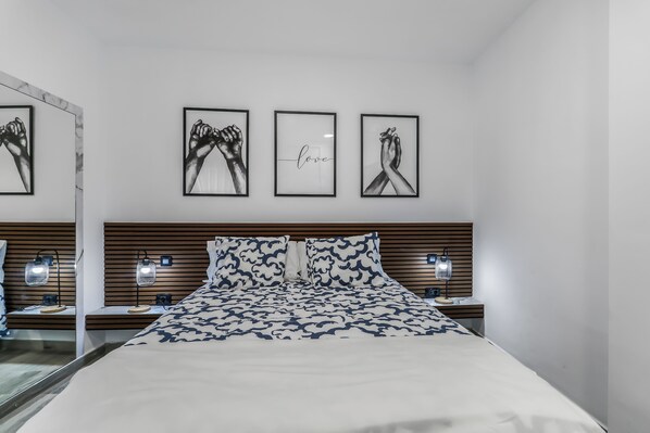 A modern bedroom with a large bed, two bedside tables with lamps, a wooden headboard, and three framed artworks on the wall.