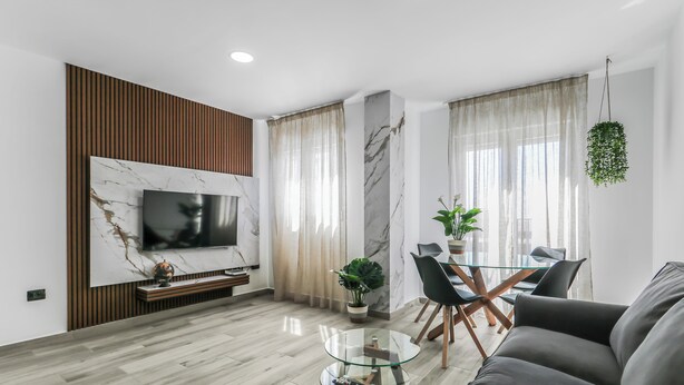 Un salon moderne avec un téléviseur à écran plat fixé au mur, une table basse en verre, un canapé, un coin repas avec une table et des chaises en verre, et une plante suspendue.