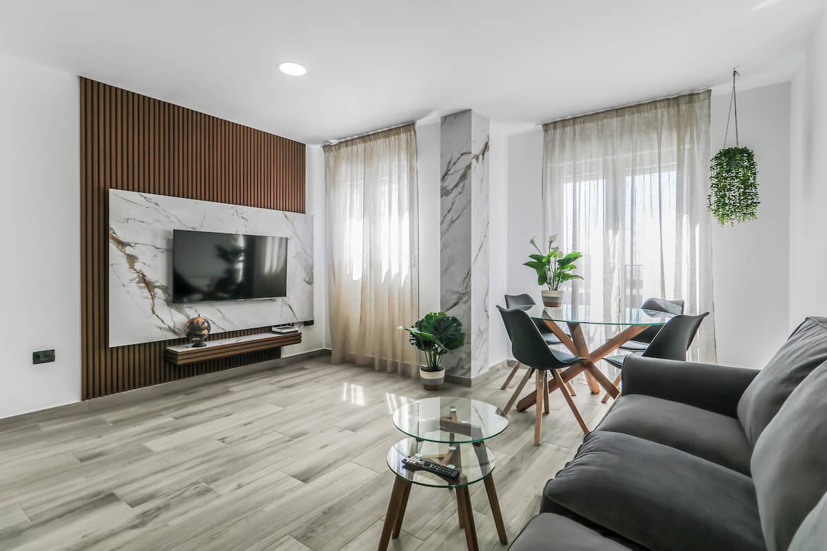 Una sala moderna con un televisor de pantalla plana montado en la pared, una mesa de centro de vidrio, un sofá, un comedor con una mesa y sillas de vidrio, y una planta colgante.