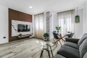 A modern living room with a flat-screen TV mounted on a wall, a glass coffee table, a sofa, a dining area with a glass table and chairs, and a hanging plant.