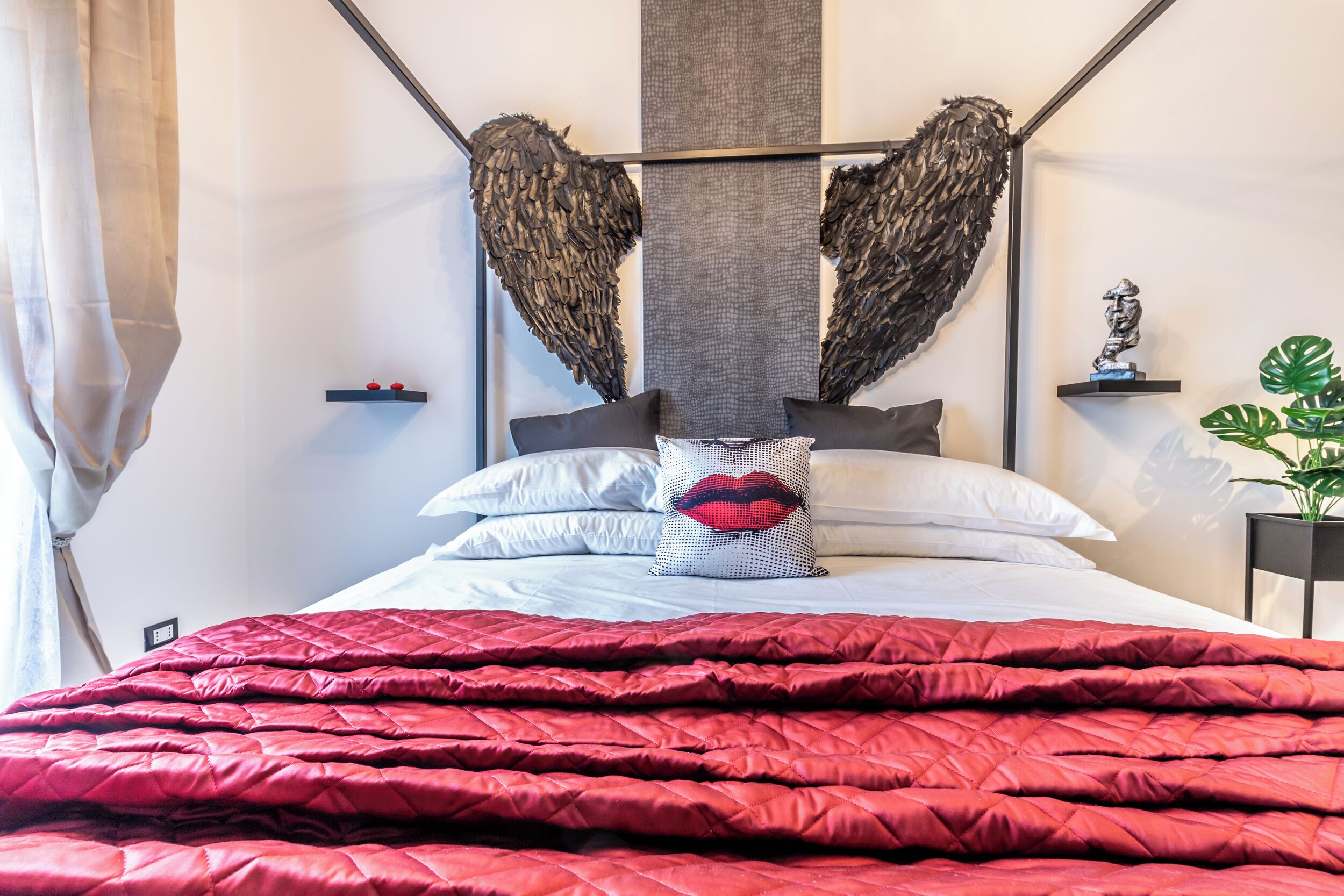 Un lit bien fait, recouvert d’une courtepointe rouge, avec un oreiller orné de motifs de lèvres rouges et une œuvre d’art murale rappelant des ailes.