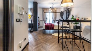 Une chambre d’hôtel moderne avec un parquet en chevrons, un lit recouvert d’une couverture rouge, un bureau avec deux tabourets, une lampe et un téléphone fixé au mur.
