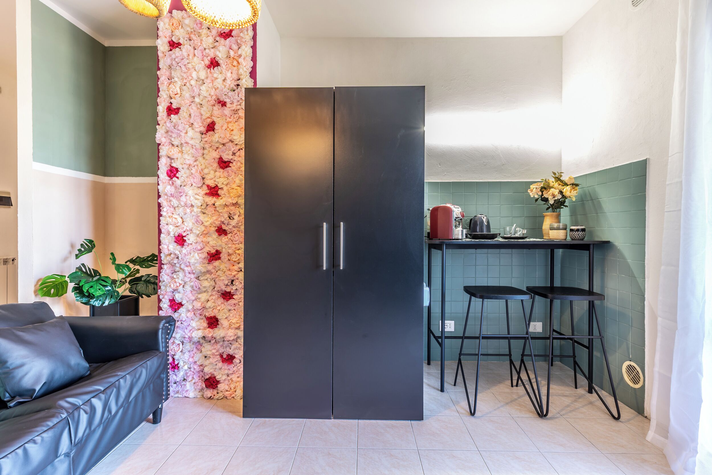 A compact living space with a black wardrobe, a sofa, a floral wall, and a kitchen area with stools.