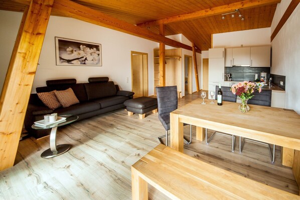 Un interior moderno con techo de madera, una zona de comedor con mesa y bancos de madera, un sofá negro con cojín, una mesita redonda con un florero y una cocina con armarios y encimera.