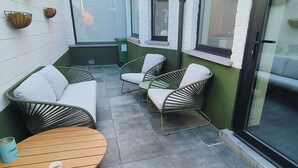 A small outdoor seating area with a round wooden table and two wicker chairs, a green trash bin, and a window with a view of the city.