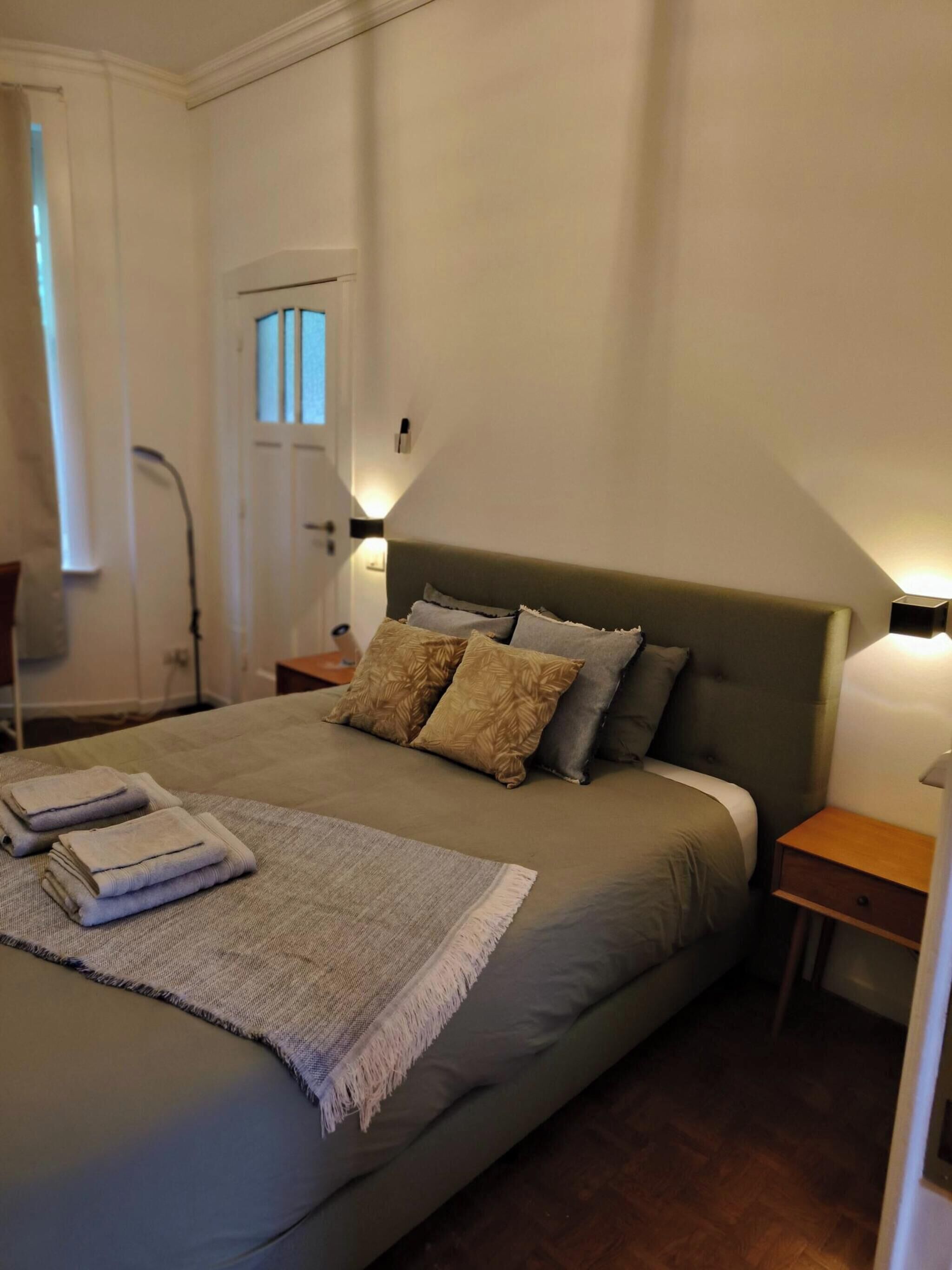 A neatly made bed with a grey bedspread, two pillows, and a folded towel. There are two bedside tables with lamps, a door, and a chair in the background.