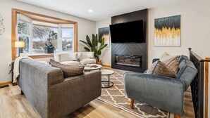 A modern living room with a fireplace, a large window, and a flat-screen TV mounted on the wall.