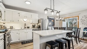 A modern kitchen with white cabinets, a large island with stools, a refrigerator, and a dining area with chairs and a table.
