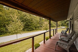 Une véranda couverte avec des balustrades en bois, une vue sur une route et deux fauteuils Adirondack.