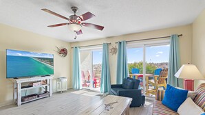 Una sala de estar con televisor, ventilador de techo, sofá y vista al exterior.