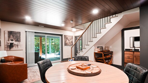 A modern living room with a round wooden table, leather chairs, a staircase, and a large window with a view of greenery. - On The Rocks | Hocking Hills, 4BR, Hot Tub (South Bloomingville)