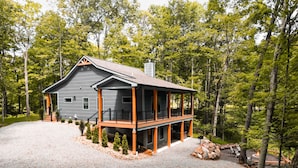 A two-story cabin with a covered porch, surrounded by trees and a gravel driveway. - On The Rocks | Hocking Hills, 4BR, Hot Tub (South Bloomingville)