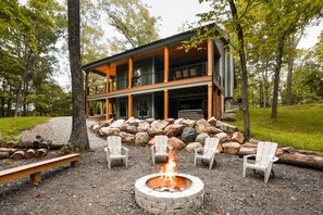 Une maison moderne à deux étages, dotée d’un foyer extérieur en pierre et de chaises blanches dans le jardin.