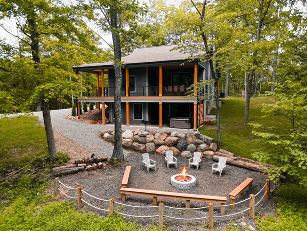 A modern two-story building with a stone fire pit and outdoor seating. - On The Rocks | Hocking Hills, 4BR, Hot Tub (South Bloomingville)