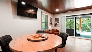 A conference room with a round wooden table, black leather chairs, a flat-screen TV, and a view of the outdoors through large sliding glass doors. - On The Rocks | Hocking Hills, 4BR, Hot Tub (South Bloomingville)