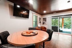 A conference room with a round wooden table, black leather chairs, a flat-screen TV, and a view of the outdoors through large sliding glass doors. - On The Rocks | Hocking Hills, 4BR, Hot Tub (South Bloomingville)