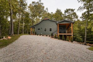 A two-story cabin with a covered porch, surrounded by a gravel driveway and forested area. - On The Rocks | Hocking Hills, 4BR, Hot Tub (South Bloomingville)