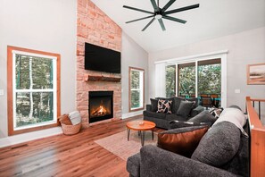 A living room with a fireplace, a sofa, a coffee table, and large windows. - On The Rocks | Hocking Hills, 4BR, Hot Tub (South Bloomingville)