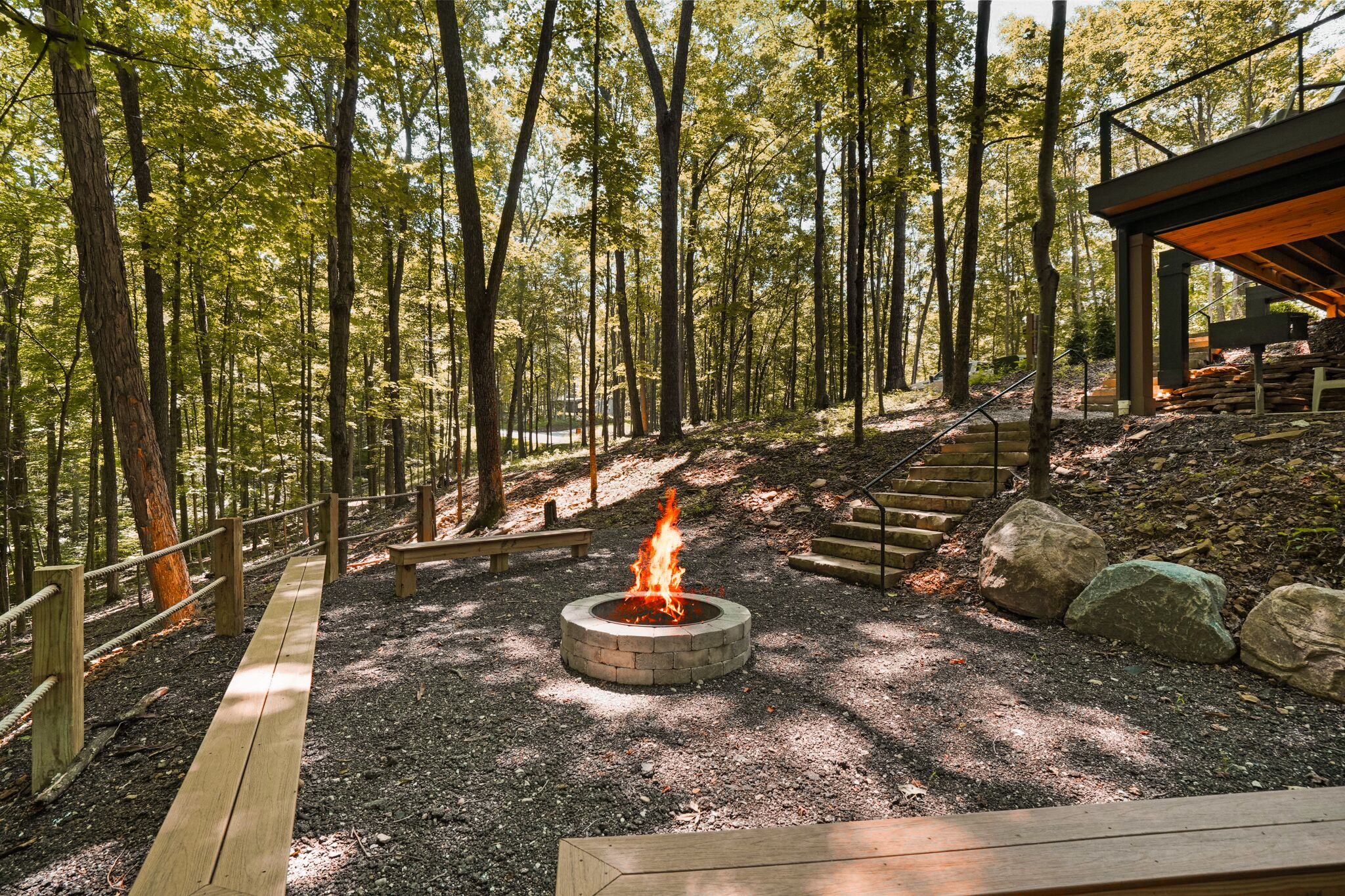 A forested area with a fire pit, wooden benches, and a covered seating area.