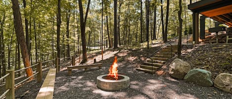 A forested area with a fire pit, wooden benches, and a covered seating area.