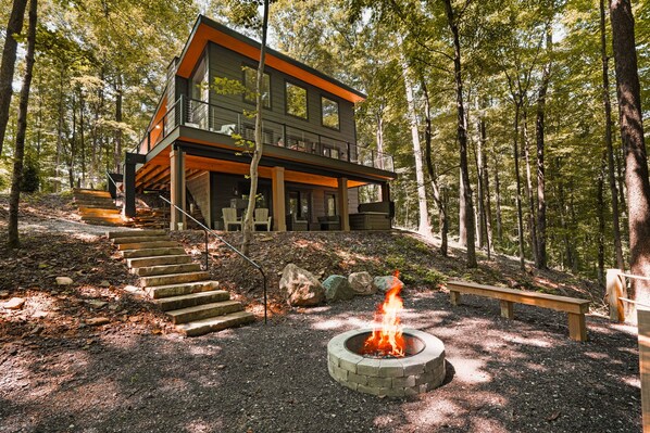 A modern two-story cabin with a fire pit in the yard, surrounded by trees.