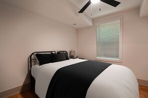 A bedroom with a bed, a ceiling fan, a window with blinds, and a nightstand with a lamp.