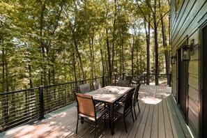 A wooden deck with a table and chairs, surrounded by a metal railing, overlooking a forested area.