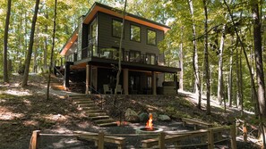 A modern two-story cabin with a deck, surrounded by a forest.