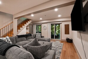 A modern living room with a staircase, grey sofas, a flat-screen TV, and a large window.