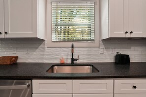 A kitchen with white cabinets, a black countertop, a stainless steel sink, a window with blinds, and a toaster.