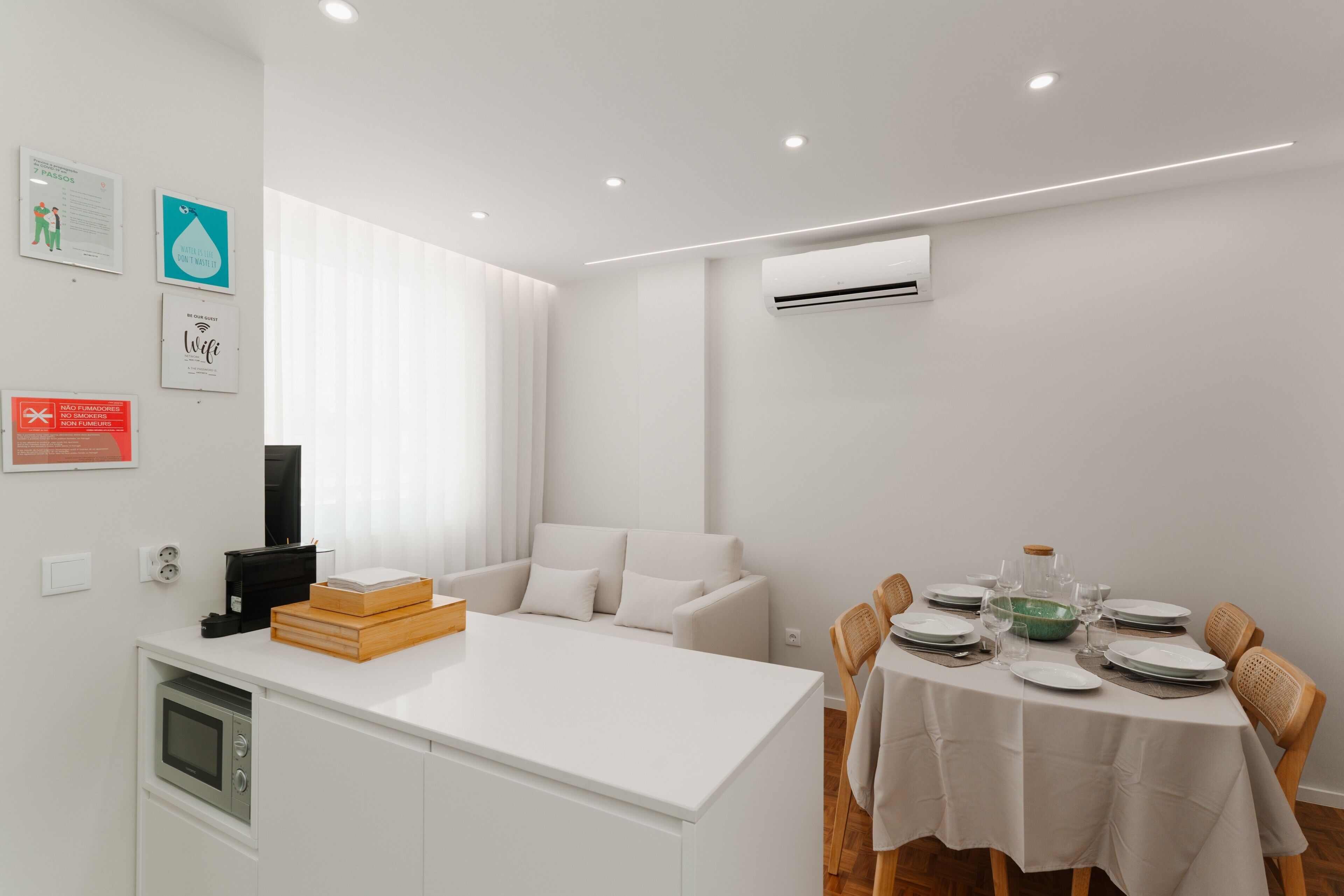 Cozinha moderna com armários brancos, área de jantar com mesa branca para quatro pessoas e um sofá branco ao fundo.