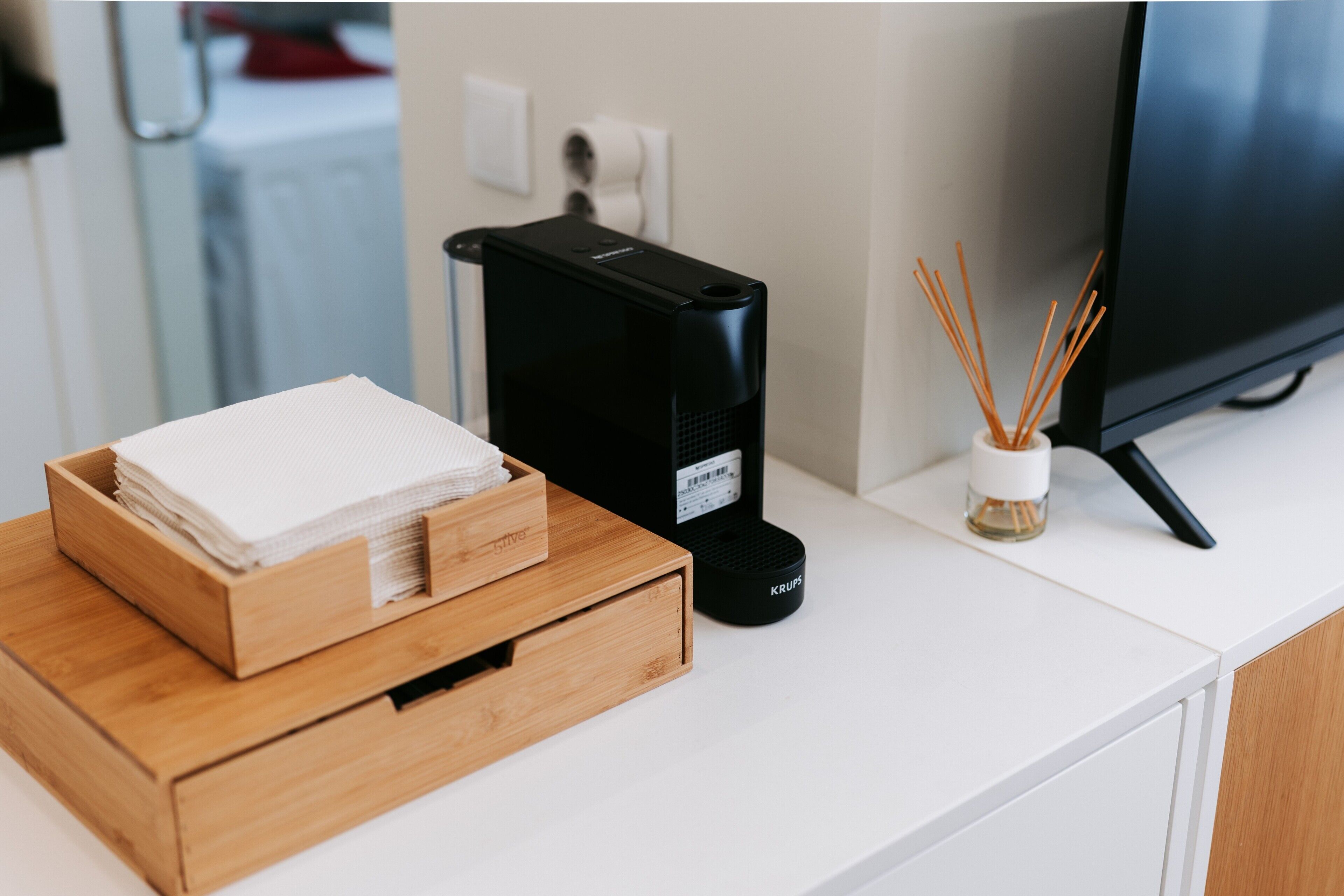 A white desk with a black coffee maker, a wooden tray with white napkins, and a small vase with reed sticks.