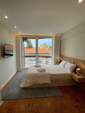 Une chambre à coucher avec un lit, un téléviseur fixé au mur, une fenêtre donnant sur des immeubles et un plancher en bois.