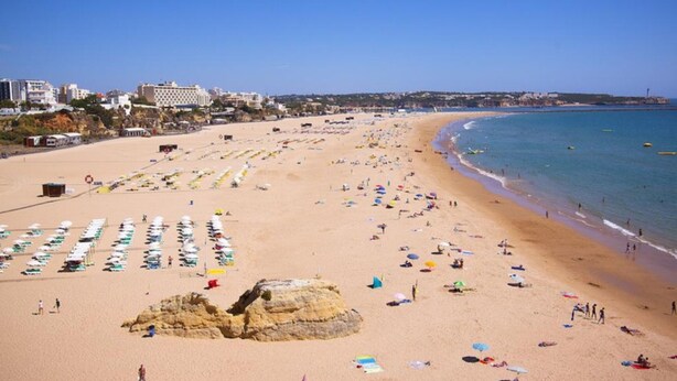 Una playa con hileras de sombrillas y tumbonas, un promontorio rocoso y edificios al fondo.