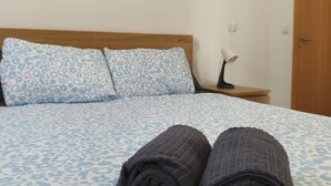 Una cama bien hecha con una colcha estampada, dos toallas enrolladas, una lámpara de noche y una puerta de madera en una habitación sencilla.