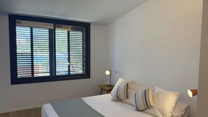 A bedroom with a bed, a window with blinds, two bedside lamps, and a bedside table.