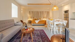 Uma sala de estar moderna com um sofá, uma mesa de centro com garrafa e copos, um tapete estampado, uma área de jantar com mesa e cadeiras, uma cama com cabeceira, e uma cozinha com eletrodomésticos.