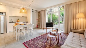 Uma sala de estar moderna com sofá, TV, área de jantar com cadeiras e mesa, e cozinha com geladeira e luminária pendente.