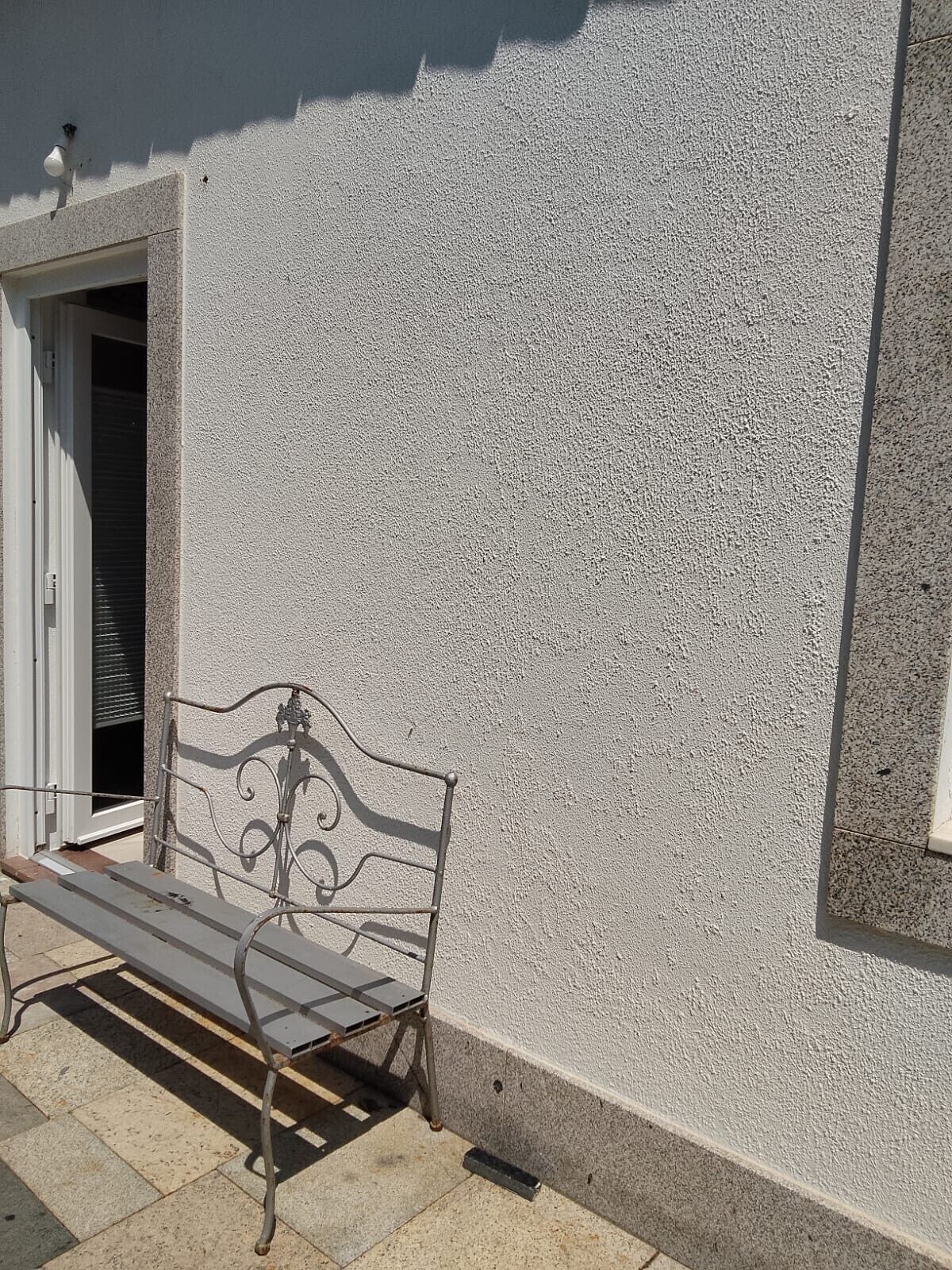 Un banc en métal adossé à un mur à texture, avec une porte et une petite fenêtre.
