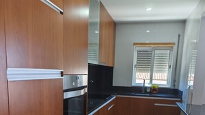 Una cocina moderna con armarios de madera, electrodomésticos de acero inoxidable y una encimera negra. Hay dos taburetes blancos y una ventana con persianas.