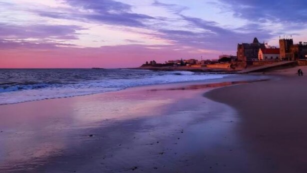 Una playa con un mar tranquilo, cielo despejado y un edificio lejano con una torre destacada.