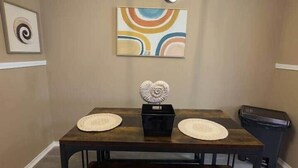 Un coin repas avec une table en bois foncé, deux sets de table ronds, un objet décoratif au centre, un luminaire moderne au-dessus et un tableau abstrait aux couleurs vives accroché au mur.
