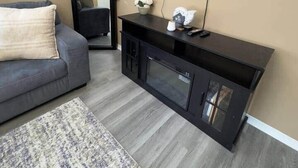 Un salon avec un canapé gris, un meuble de rangement noir pour appareils électroniques, un téléviseur à écran plat et un tapis à motifs.