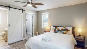 A bedroom with a bed, a ceiling fan, a nightstand with a lamp, a door, and a window.
